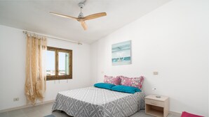 Une chambre à coucher avec un lit, une table de chevet, un ventilateur de plafond, une fenêtre avec des rideaux et un tableau au mur.