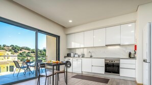 A modern kitchen with white cabinets, a dining area with a table and chairs, and a large sliding glass door leading to a balcony with a view of a hillside.