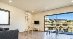 Un salón moderno con un sofá gris, una pequeña mesa con una planta, un televisor y un balcón con una mesa y sillas de comedor.
