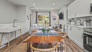 Una cocina moderna con armarios blancos, horno empotrado y comedor con mesa y sillas de madera.