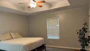 Un dormitorio con cama, ventilador de techo, ventana con persianas y planta en maceta sobre una mesa.