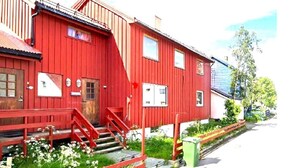 Punainen puinen rakennus, jossa on portaikko, joka johtaa sisäänkäyntiin, pieni puutarha-alue, jossa on kukkia, ja vihreä roskakori.
