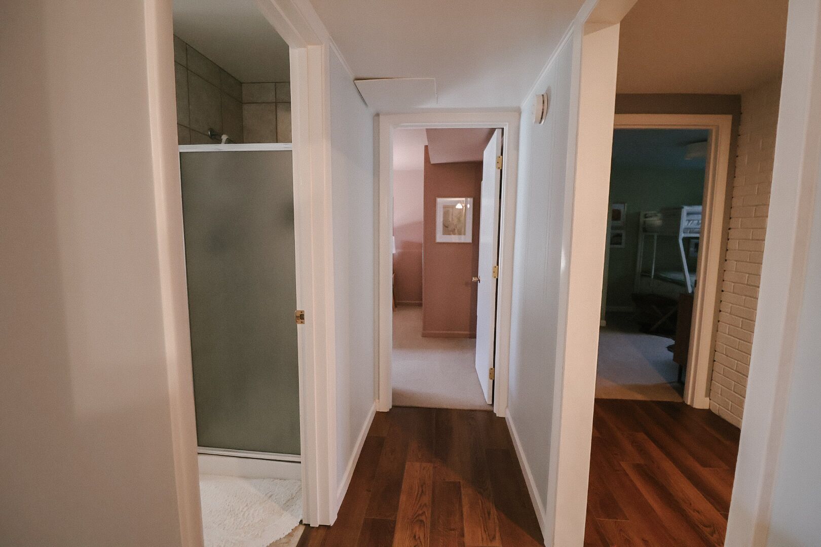 A hallway with wooden flooring, white walls, and a glass door leading to a bathroom.