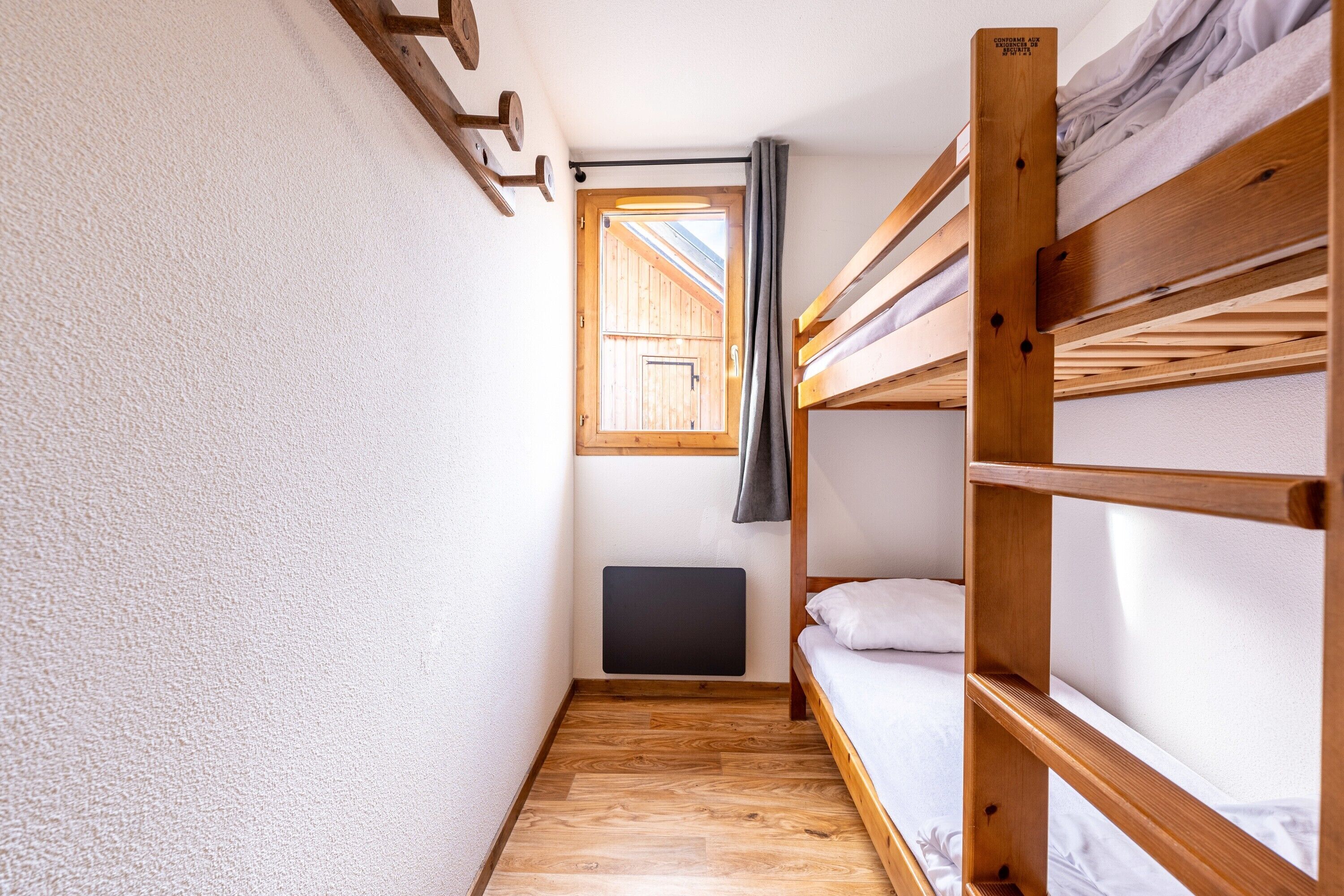 A narrow room with wooden bunk beds, a window with curtains, and a radiator on the wall.
