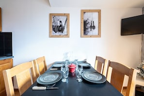 A dining table set with plates, glasses, and cutlery, flanked by wooden chairs, with a TV mounted on the wall above.