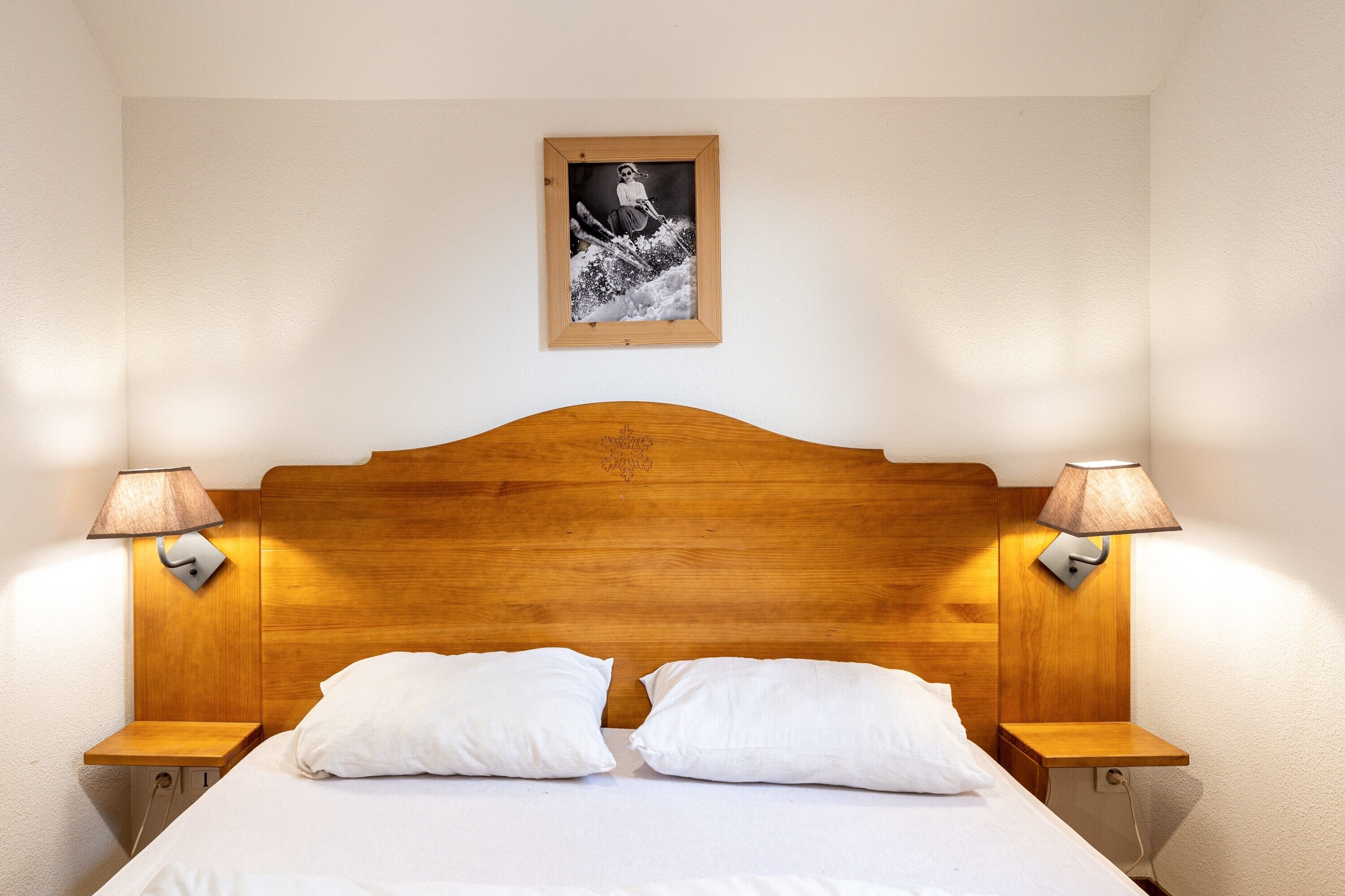 Een net opgemaakt bed met twee kussens, een houten hoofdbord en twee bedlampjes.