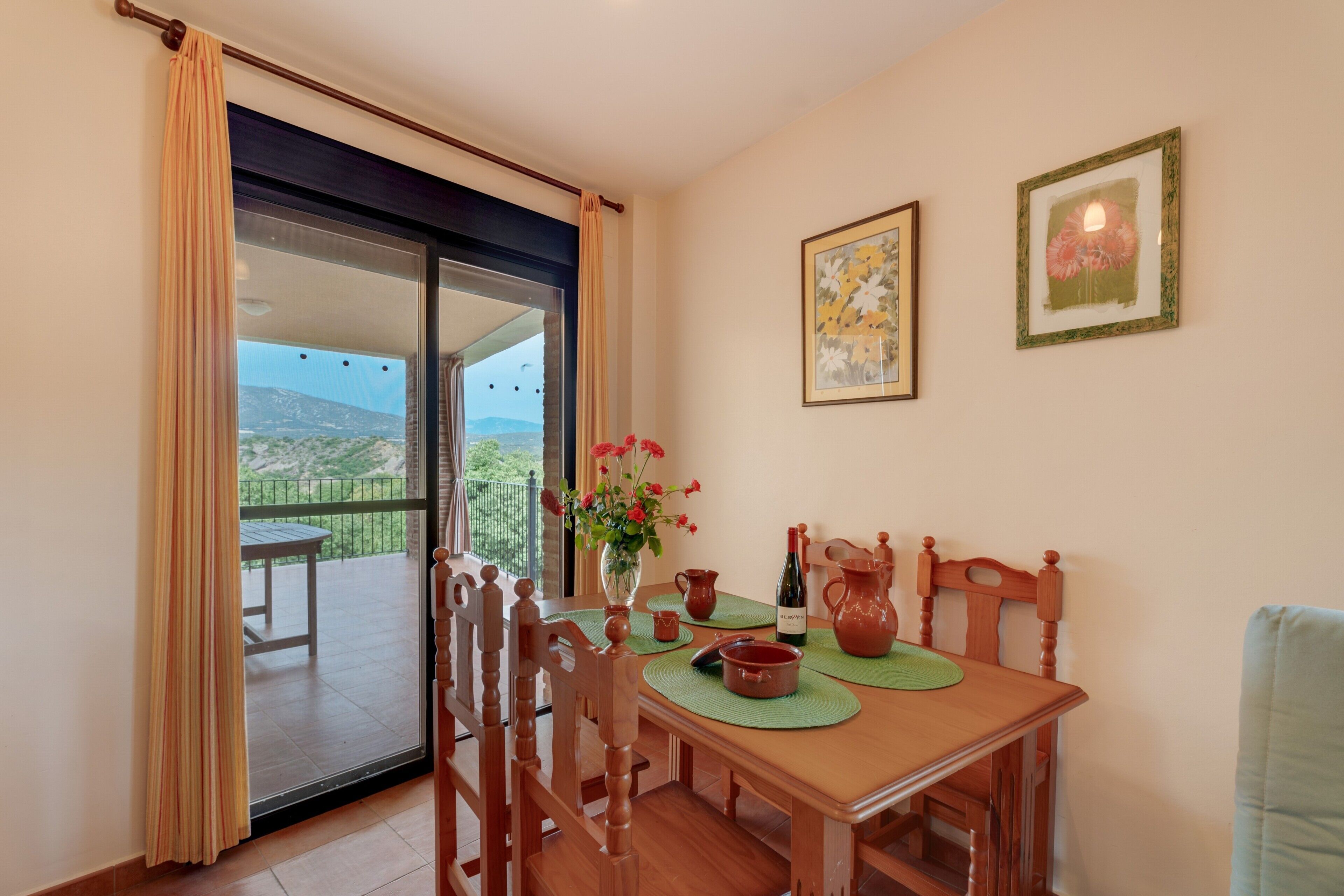 Uma sala de jantar com uma mesa, cadeiras, um vaso com flores, uma garrafa de vinho e vista para uma varanda com mesa e cadeiras.