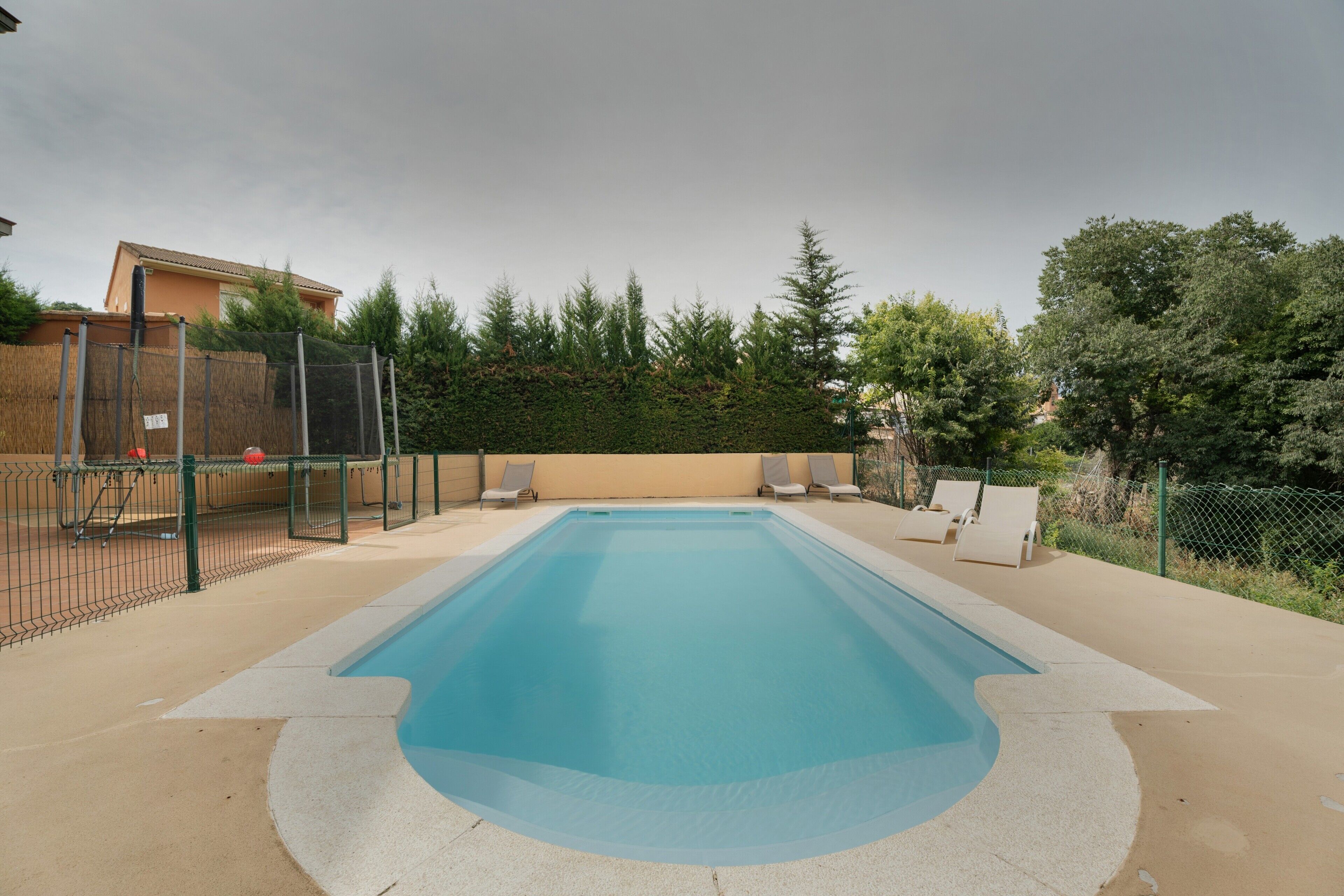 A rectangular swimming pool with a surrounding deck, a fence, and a few lounge chairs.