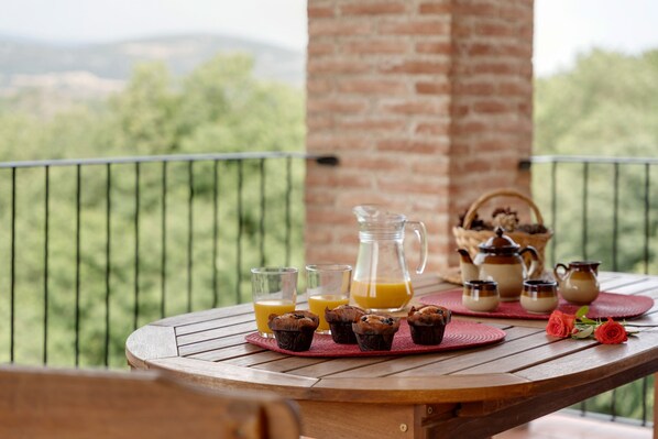 A wooden table with a pitcher, glasses, muffins, and a basket on a balcony with a view of greenery.