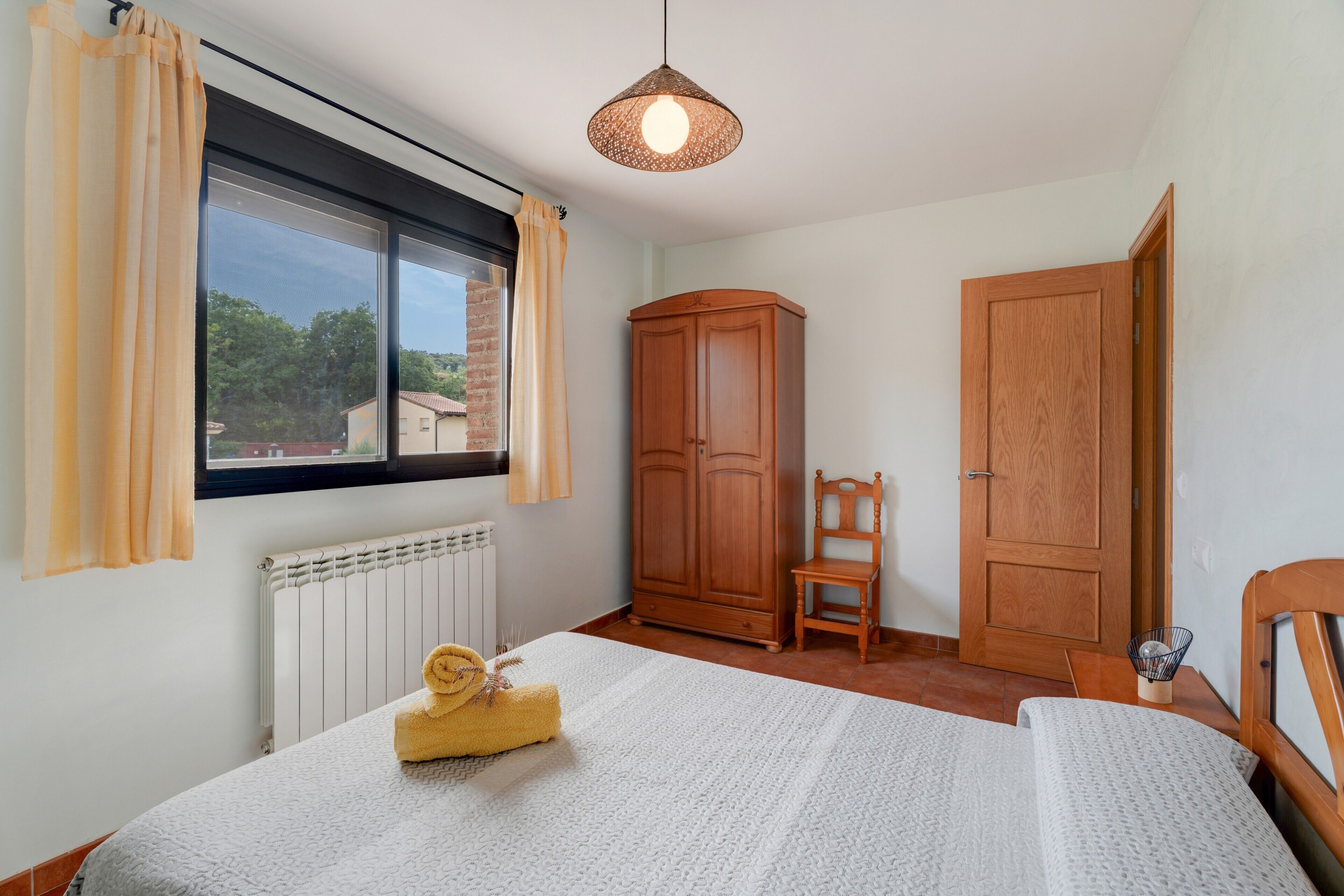 Un dormitorio con cama, un radiador, una ventana con cortinas, un armario, una silla y una lámpara colgante.