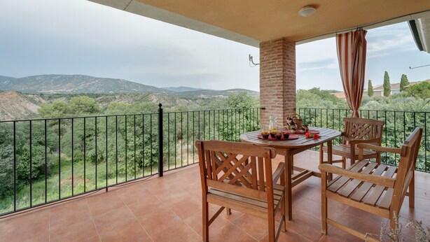 A balcony with a table and chairs, a brick pillar, and a view of green hills. - Holiday Home 'Sueños De Guara Casa Rural' with Mountain Views, Private Pool and Wi-Fi (Casbas de Huesca)