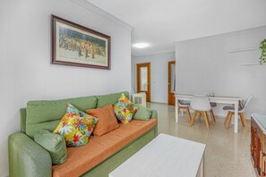 A living room with a green sofa, colorful cushions, a white coffee table, a dining area with a white table and chairs, and a framed picture on the wall. - Divina Pastora Sanlucar Apartment with Balcony and Air Conditioning (Sanlúcar de Barrameda)