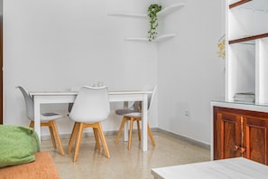 Une pièce avec une table blanche, deux chaises, un coussin vert, un meuble en bois et une plante suspendue.
