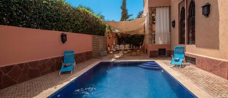 Una piscina con dos sillas reclinables azules, un porche cubierto y un edificio con fachada color beige y ventanas arqueadas.