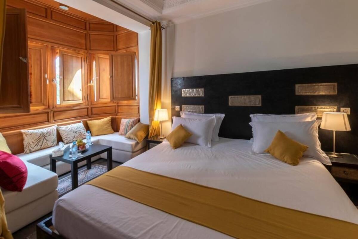 Habitación de hotel con una cama grande, un sofá y una mesita. Se observan armarios de madera y un cabecero negro.