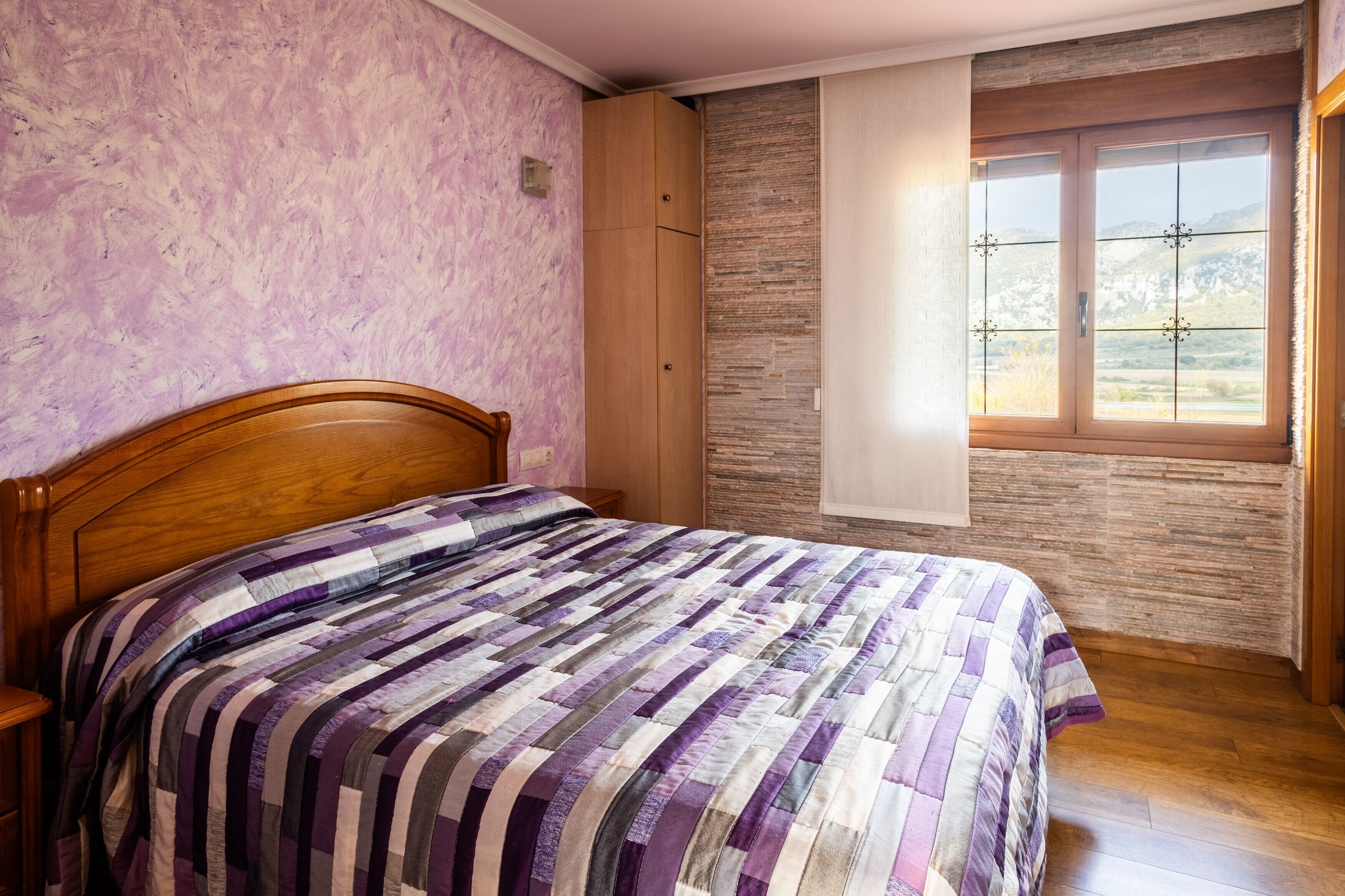 A bedroom with a wooden bed, a stone wall, and a window with a view of mountains.