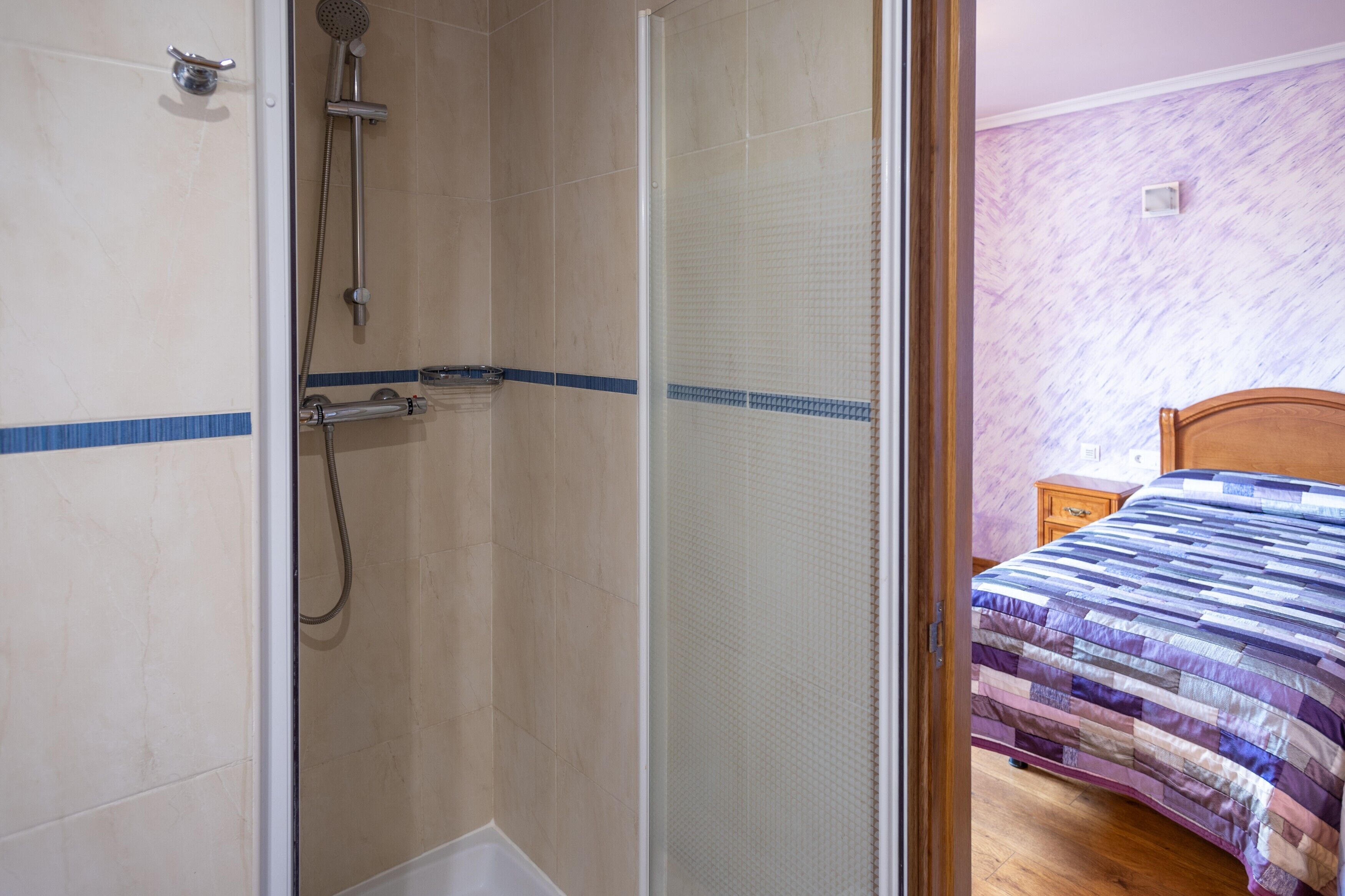 Un baño con ducha, una cama con colcha y una mesita de noche de madera.