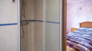 Un baño con ducha, una cama con colcha y una mesita de noche de madera.