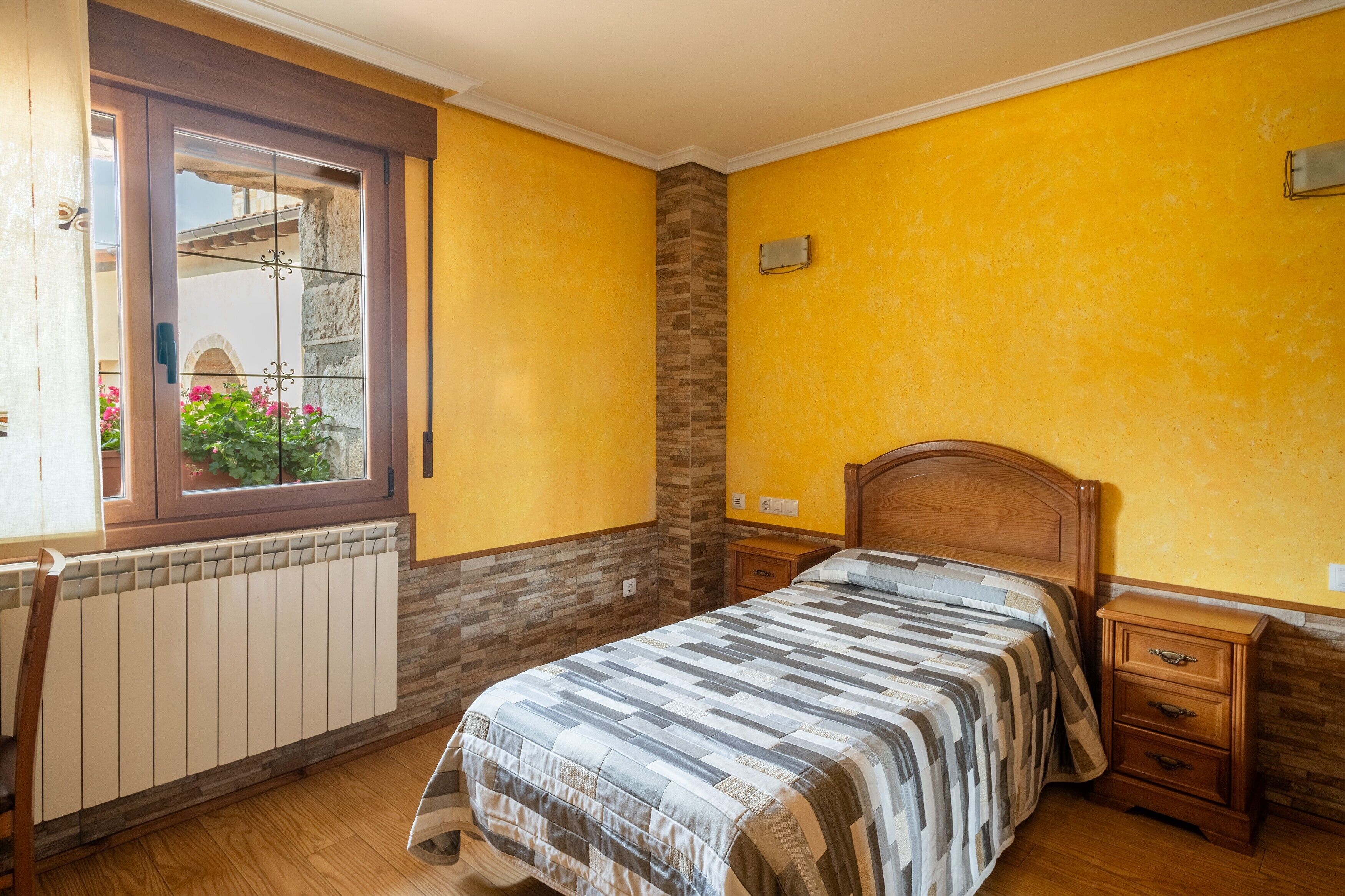 Un dormitorio con cama de madera, una mesita de noche, una ventana con vistas a flores y un radiador.