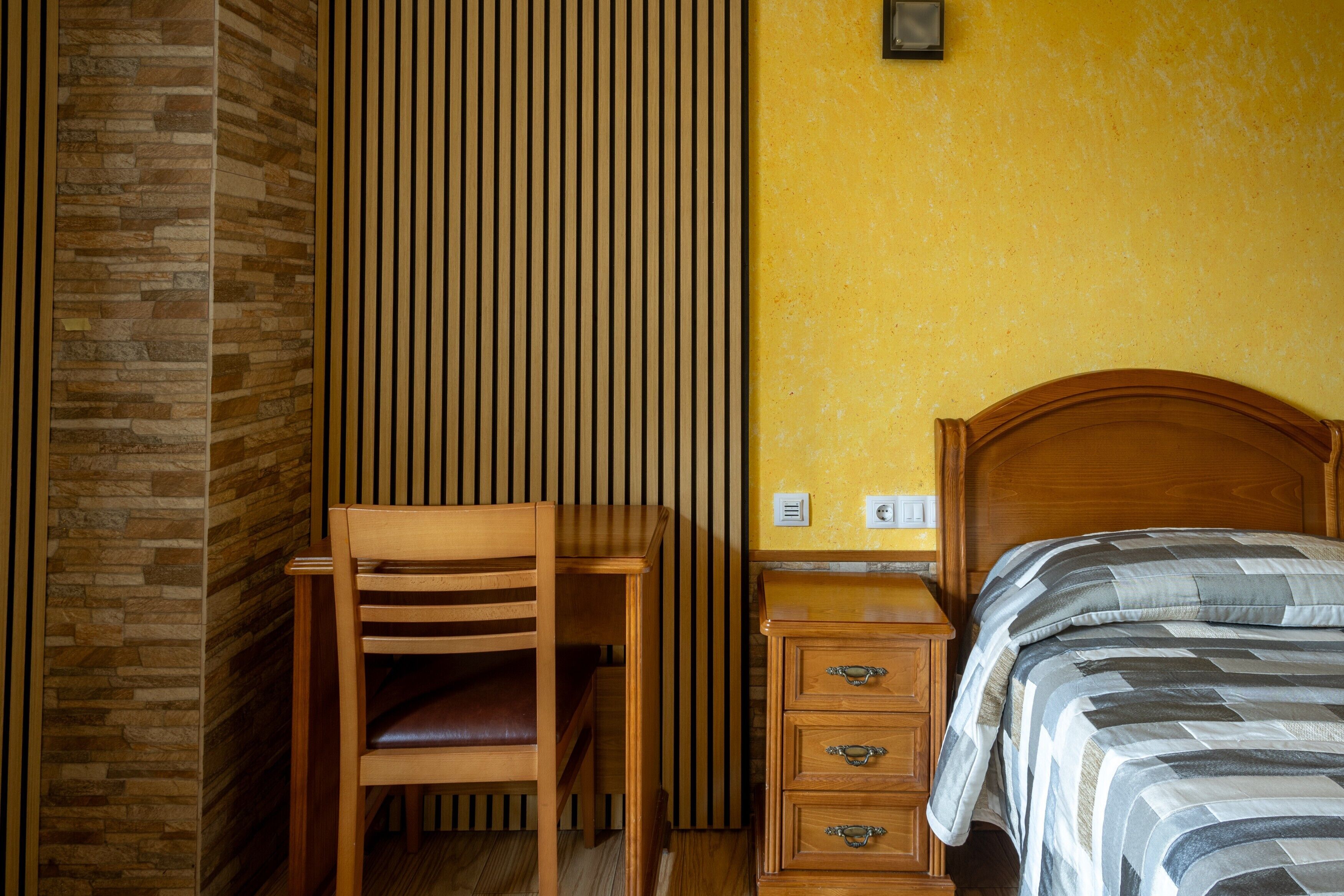 Een kamer met een houten bureau, een stoel, een bed met gestreept beddengoed, een nachtkastje en een wand met verticale jaloezieën.