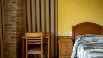 Een kamer met een houten bureau, een stoel, een bed met gestreept beddengoed, een nachtkastje en een wand met verticale jaloezieën.