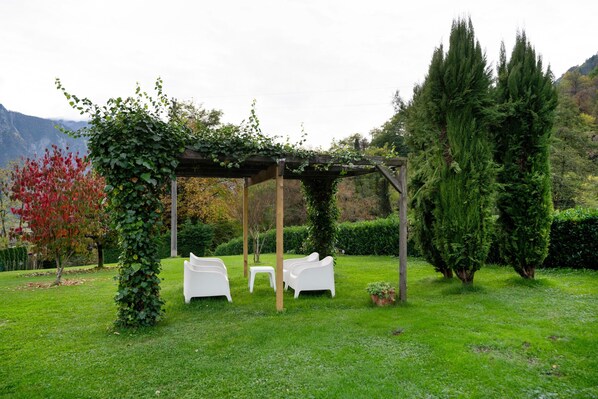 A gazebo with white chairs and a table, surrounded by greenery and trees.