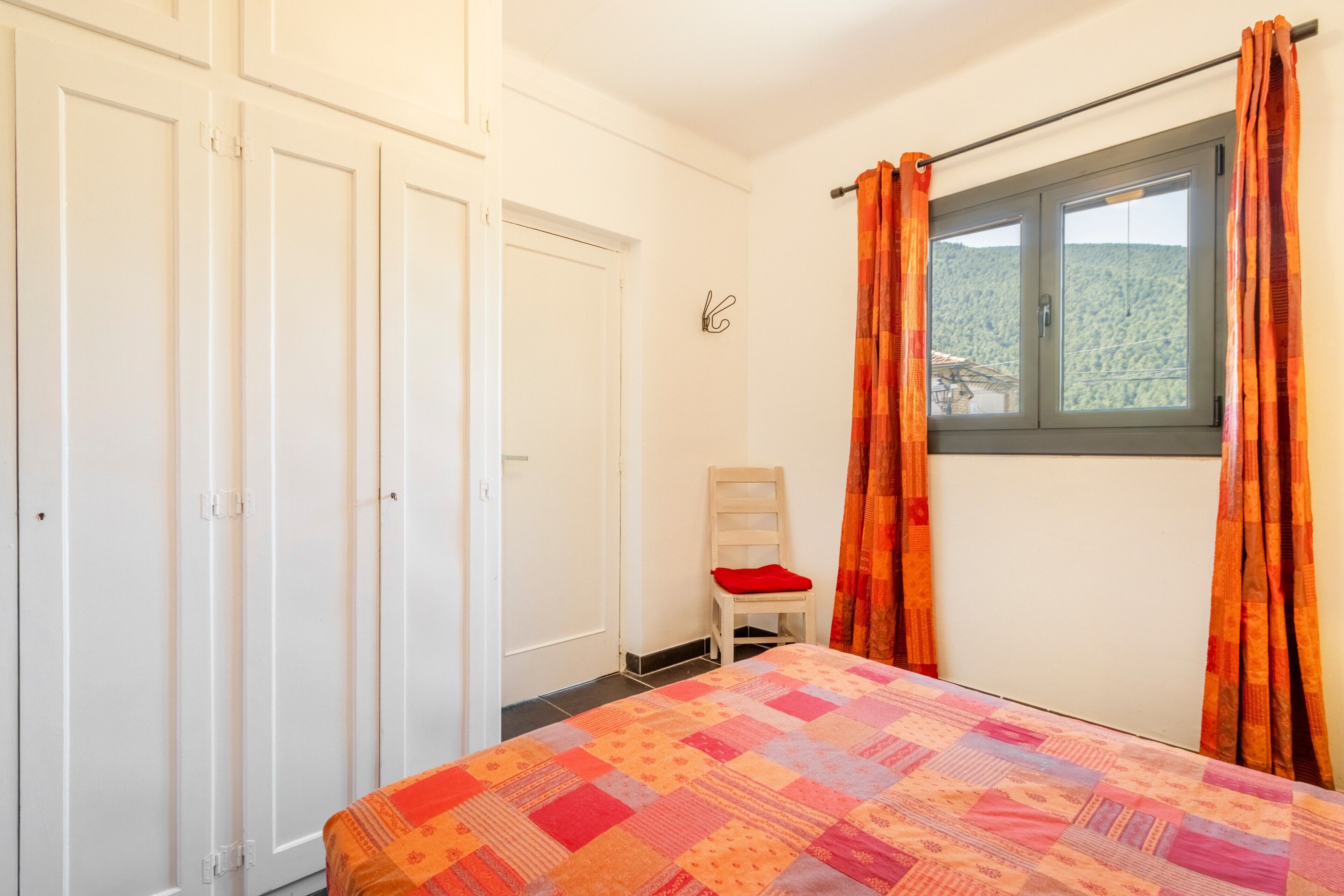 A bedroom with a bed, a chair, a window with curtains, and a wardrobe.