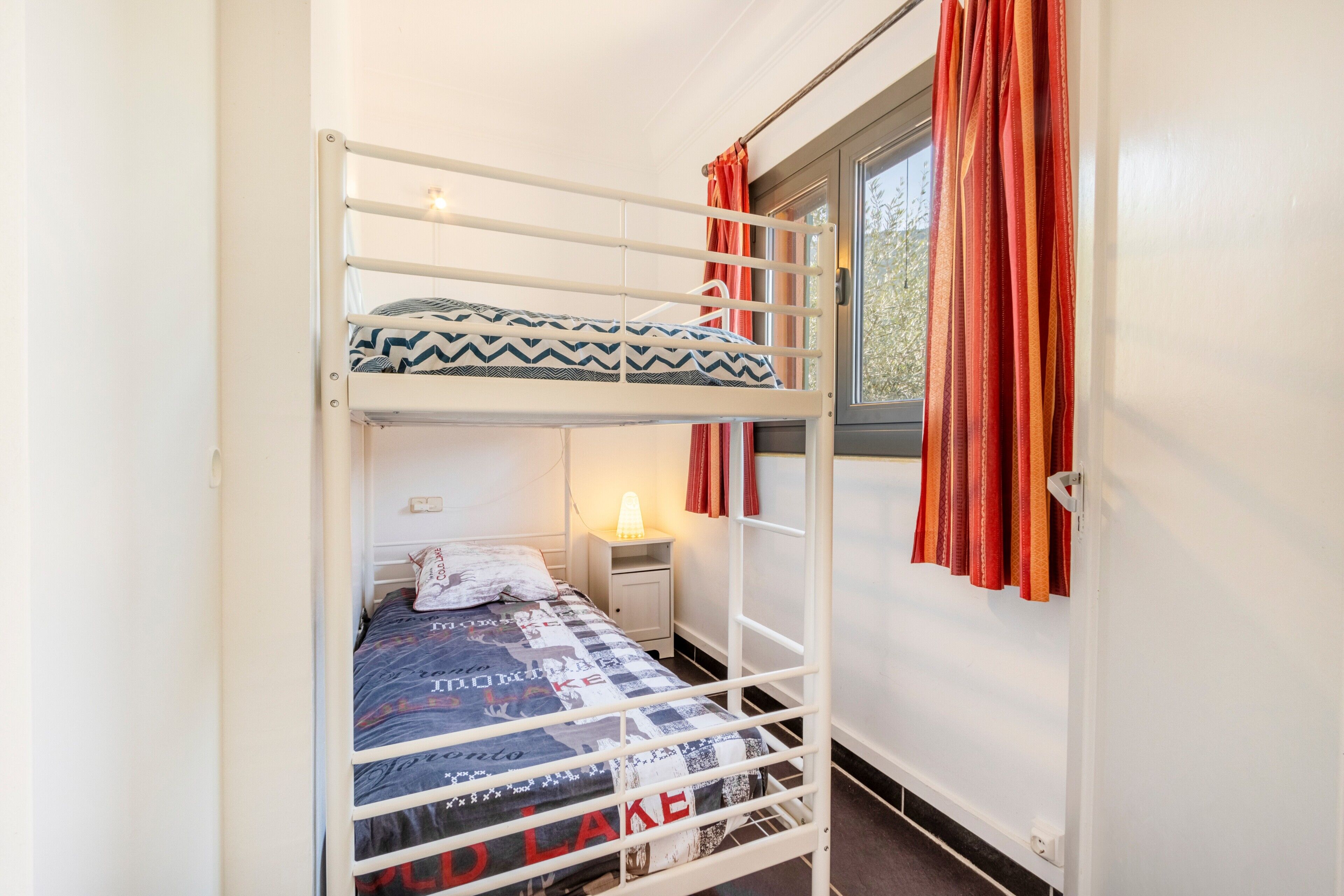 A bunk bed room with a window, curtains, a bedside table, and a quilted bedspread.