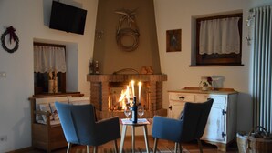 A cozy living room with a fireplace, two chairs, a small table, and a television mounted on the wall. - Chalet 'Baita Casapendola' (Roncegno Terme)