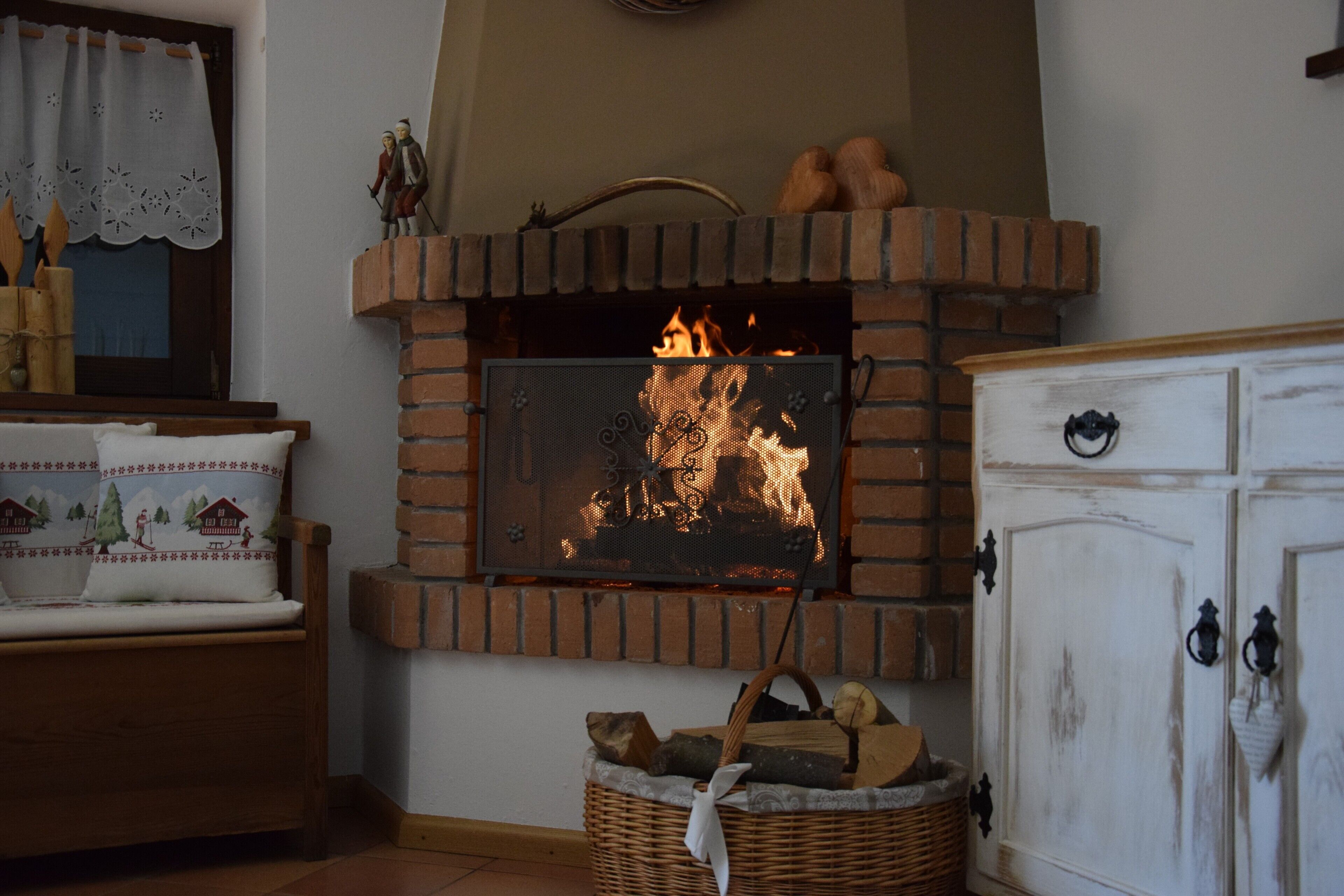 Une cheminée avec un feu, un banc en bois, un panier en osier rempli de bûches et un meuble blanc avec des poignées noires.