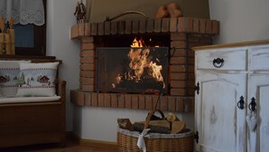 A fireplace with a fire, a wooden bench, a wicker basket with logs, and a white cabinet with black handles. - Chalet 'Baita Casapendola' (Roncegno Terme)