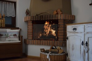 Une cheminée avec un feu, un banc en bois, un panier en osier rempli de bûches et un meuble blanc avec des poignées noires.