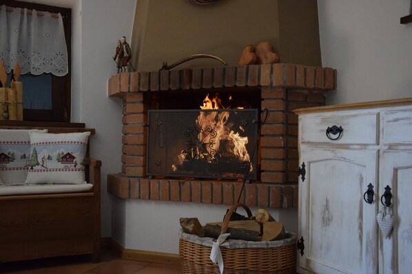 Ein Kamin mit Feuer, eine hölzerne Bank, ein Korbkiste mit Holzscheiten und ein weißes Schränkchen mit schwarzen Griffen.