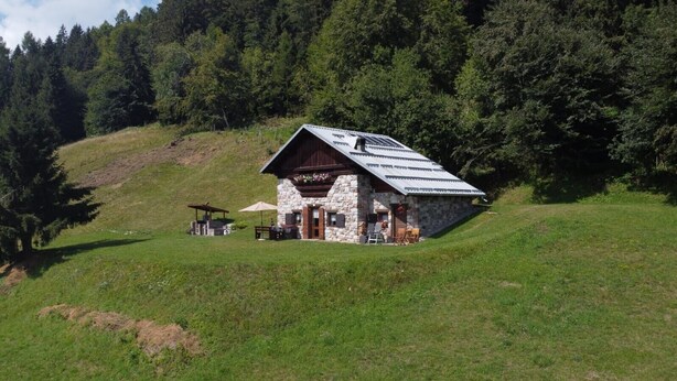 A stone cabin with a sloped roof, a patio area with outdoor seating, and a view of a forested hillside. - Chalet 'Baita Casapendola' (Roncegno Terme)