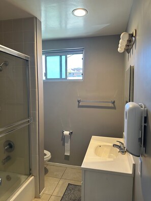 Bathroom features a clear shower area, toilet, and sink, with a window and hand sanitizer dispenser.
