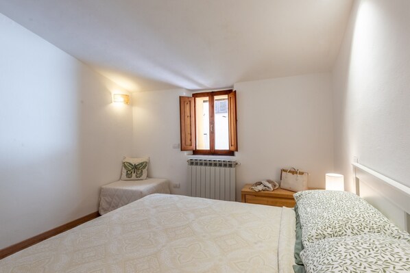 Ein Schlafzimmer mit Bett, Nachttisch, Lampe, einem Fenster mit Holzjalousien und einer Wandleuchte.