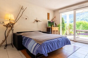 A bedroom with a bed, a lamp, a TV, and a view of greenery through a sliding door. - Holiday Apartment "T3 Au Vert À 5 Min D'Annecy Centre" with Private Terrace, Private Garden & Wi-Fi (Annecy)