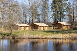 Three cabins with wooden exteriors, a picnic table, and a pond in the foreground. - Charming Safari Tent by the Lake (Gasselternijveen)