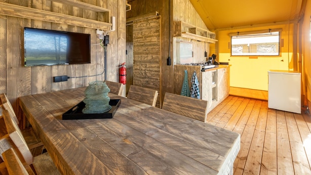 A cozy cabin interior with a wooden dining table, a kitchen area, and a television mounted on the wall. - Charming Safari Tent by the Lake (Gasselternijveen)