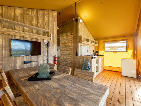 A cozy cabin interior with a wooden dining table, a kitchen area, and a television mounted on the wall. - Charming Safari Tent by the Lake (Gasselternijveen)