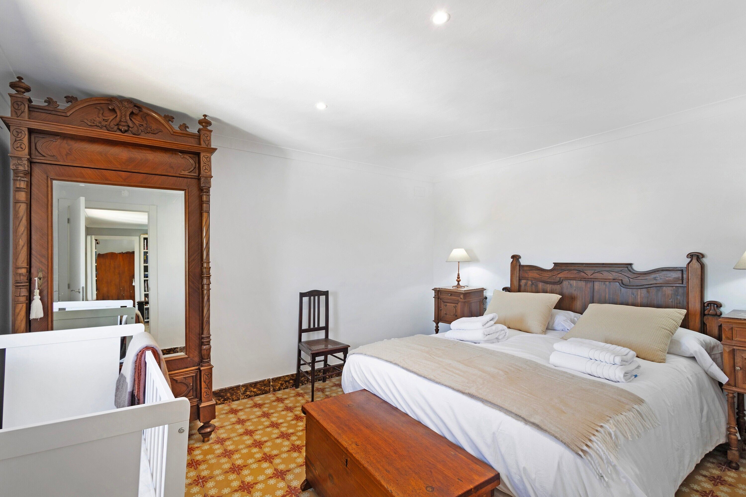 A bedroom with a wooden bed, a wooden nightstand, a bedside table, a chair, a wooden bench, a mirror, and a patterned floor.