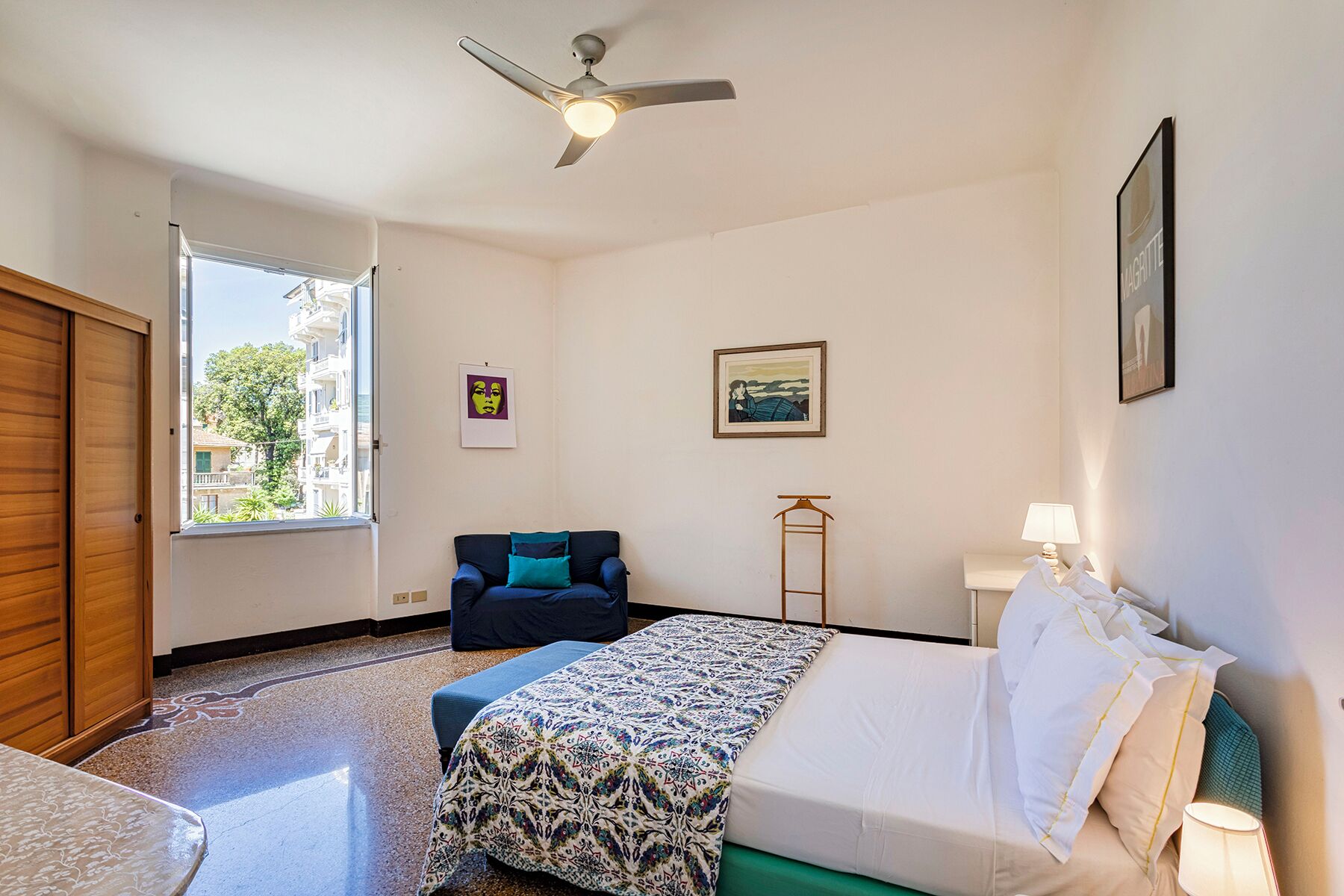 Une chambre d’hôtel avec un lit, une chaise, une petite table, un ventilateur de plafond et une fenêtre donnant sur des immeubles et des arbres.