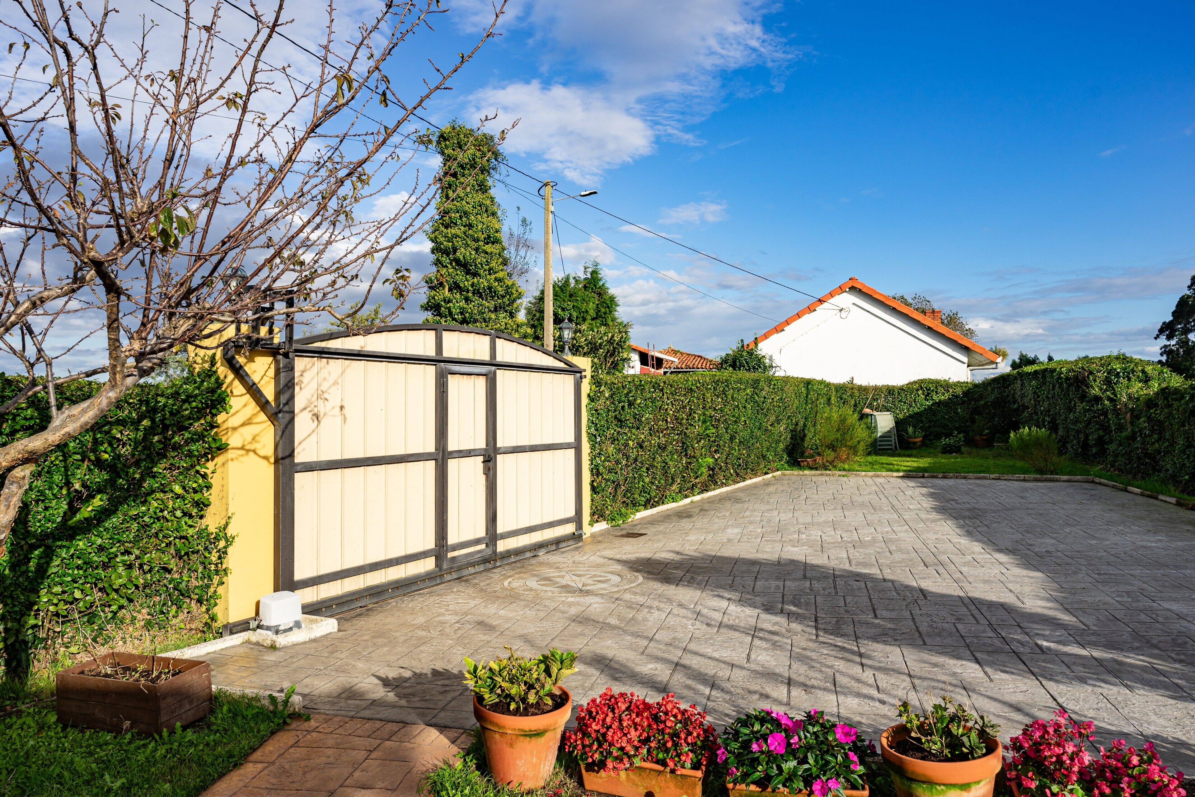 A paved driveway leading to a gated entrance, surrounded by greenery and potted plants.