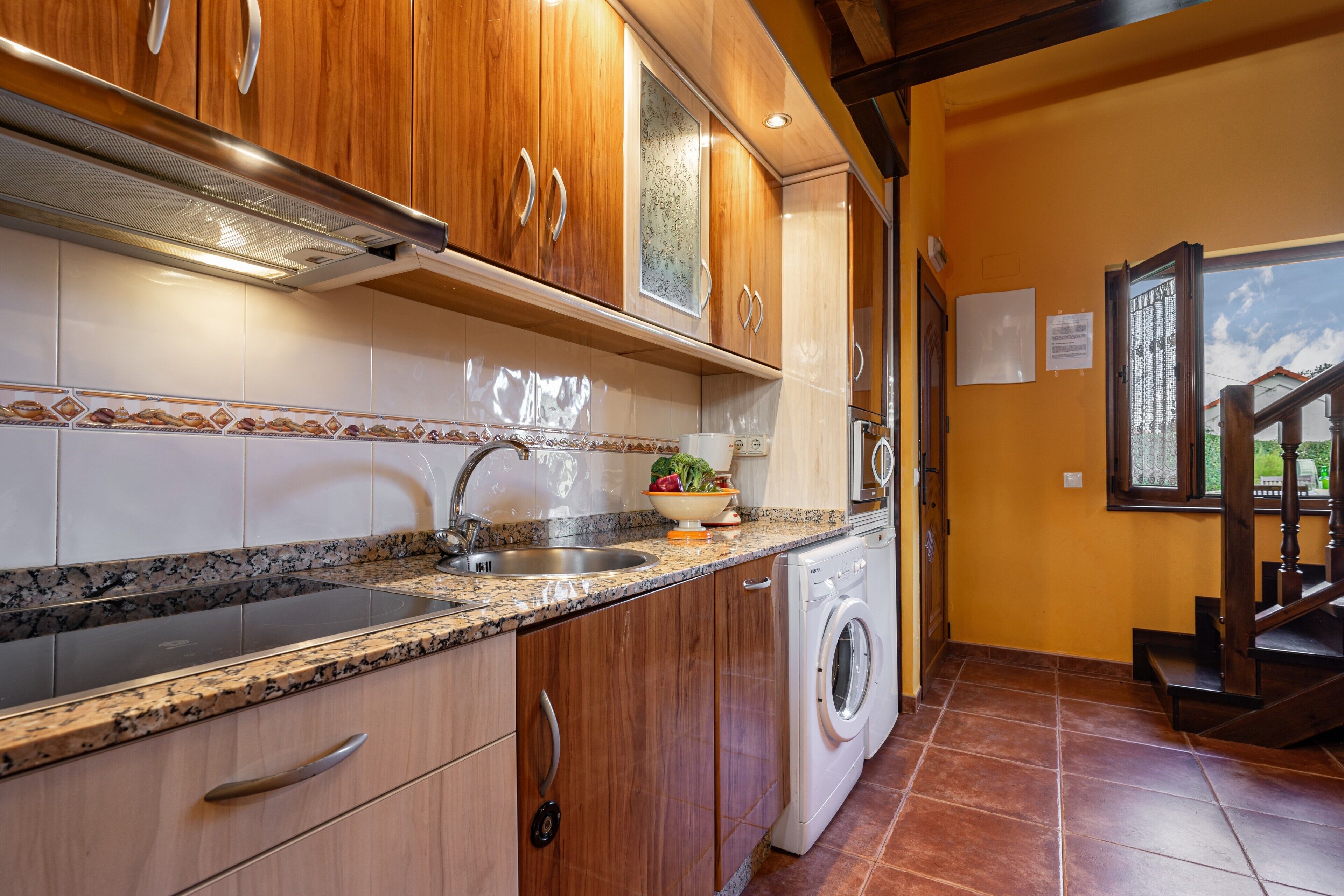Una cocina con gabinetes de madera, un lavamanos de acero inoxidable, una lavadora y un salpicadero de azulejos.
