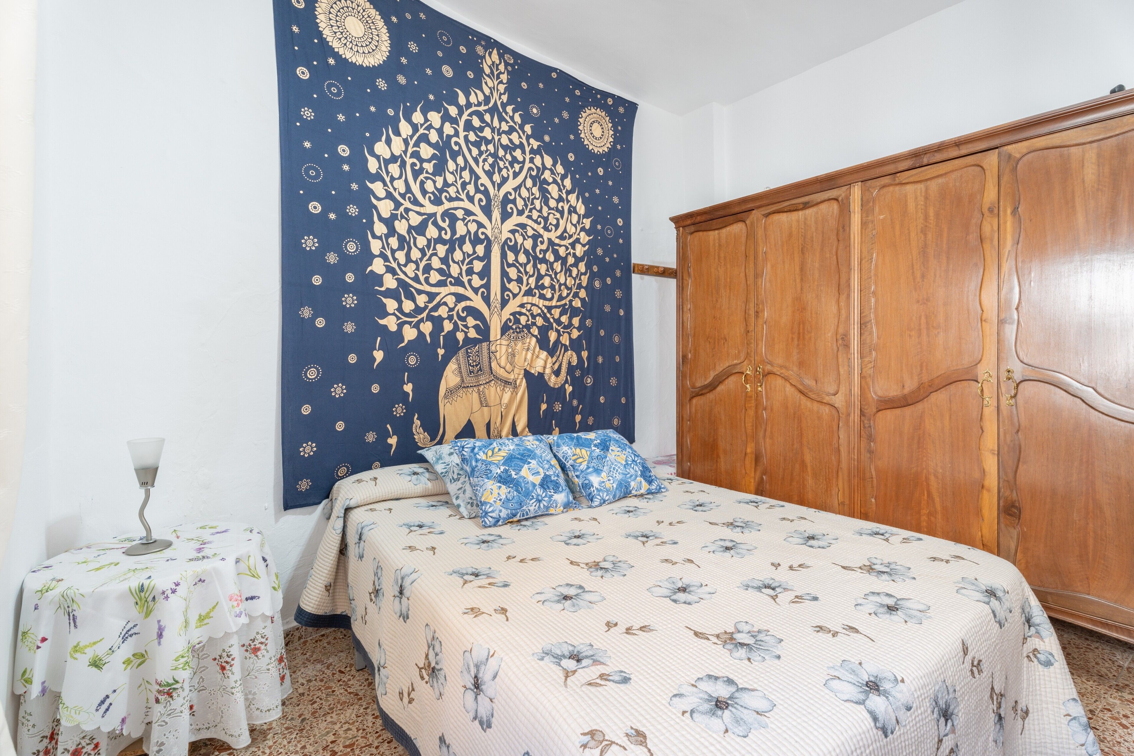 Une chambre avec une grande armoire, un lit avec une literie à motifs floraux, une tapisserie représentant un arbre et une petite table avec une lampe.