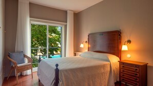 Un dormitorio con un ventanal grande, una cama con cabecera de madera, una silla de mimbre y una mesita de noche con una lámpara.