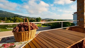 Un balcón con una cesta de mimbre llena de uvas, una mesa de madera y vistas a un paisaje verde con casas y un puente.