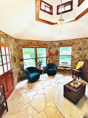 A spacious living area with stone walls, two blue armchairs, a wooden coffee table, and a ceiling fan.