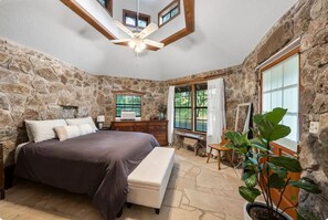 A bedroom with a stone wall, a bed, a bench, a desk, and a potted plant.