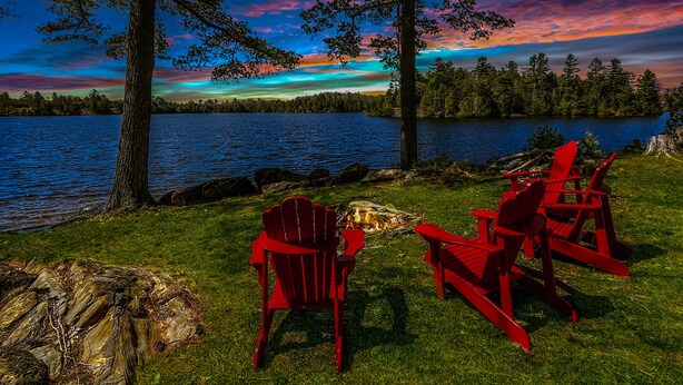 A lakeside view with red chairs and a campfire.